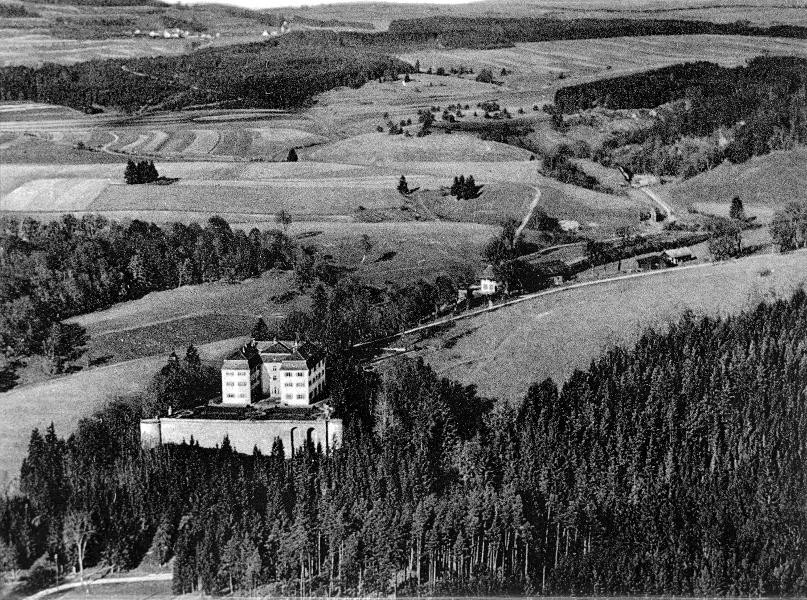 Historische Luftaufnahme des Schlosses und des gesamten Geländes in Grafeneck 1930. Im Vordergrund befindet sich das Schloss auf einer Anhöhe, umgeben von Wald. Im Hintergrund befindet sich das landwirtschaftliche Hauptgebäude und am rechten Bildrand das Gebäude, welches 1940 zur Gaskammer umgebaut wurde. Dahinter sind Felder und Bäume
