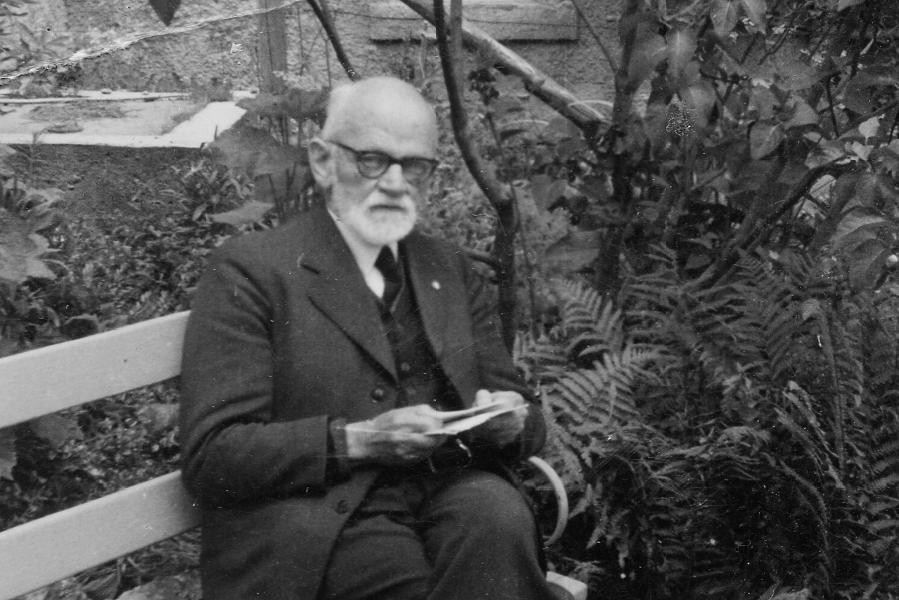 Heinrich Hermann auf einer Bank im Freien. Das Bild zeigt einen älteren Mann mit schwarzer Brille und weißem Vollbart. Er trägt einen Anzug und sitzt auf einer Bank. Die Beine hat er übereinander geschlagen. In den Händen hält er mehrere Blätter Papier. Er blickt ernst in die Kamera. Im Hintergrund sind eine Hauswand und unterschiedliche Pflanzen zu sehen.